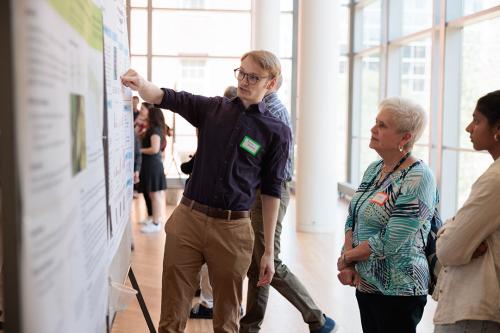 A Biology student stands in front of a large research poster and points out parts of it to two other people