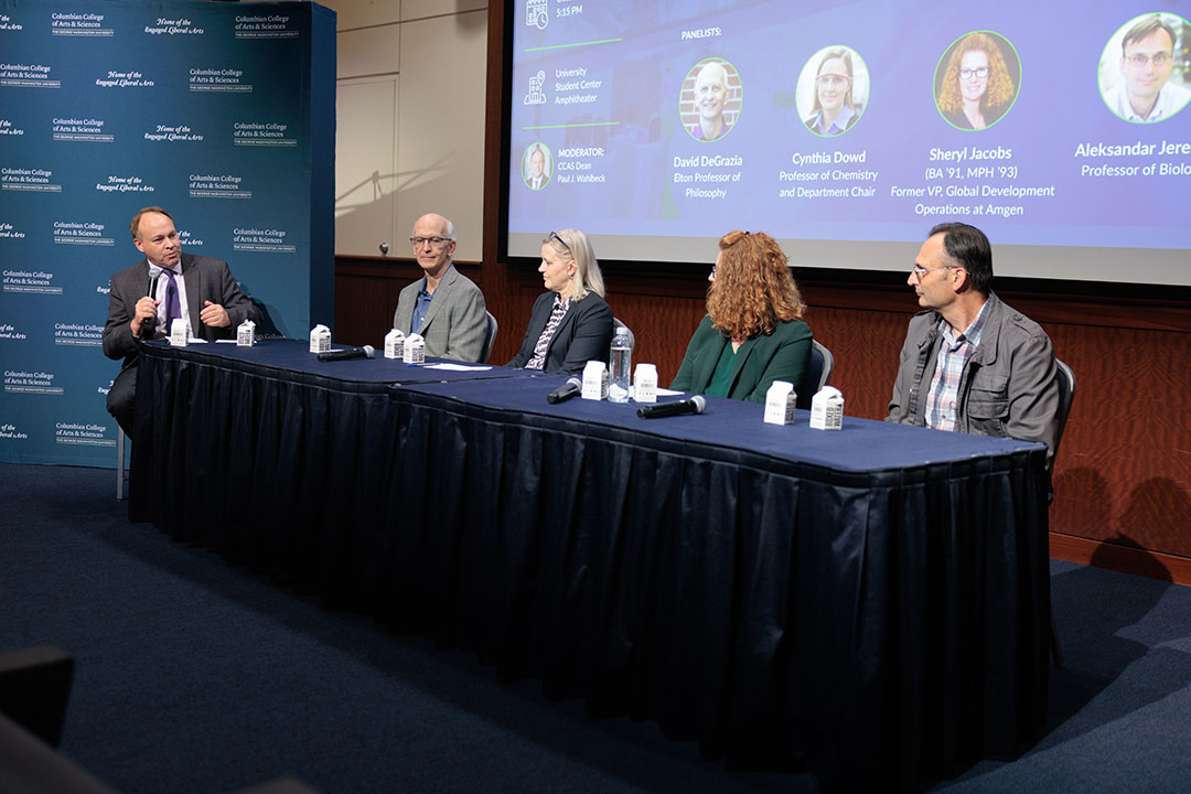 Dean Paul Wahlbeck moderating a panel discussion with three faculty members and a GW alum seated behind a draped table on stage